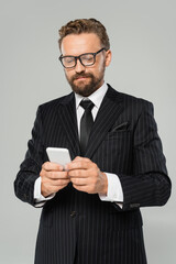bearded businessman in glasses and suit typing on smartphone isolated on grey .