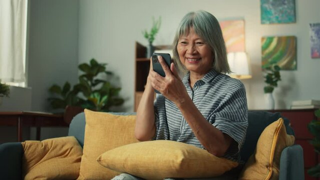 Happy Asian elderly woman smile holding mobile phone checking email received good news, feeling surprised excited. Cheerful aged female looking at smartphone screen sit on sofa in home living room.