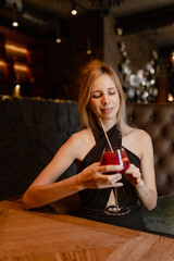  Beautiful young woman with loose hair in evening black dress with glass of alcohol is sitting at bar against background of luxurious interior of expensive restaurant. Atmosphere of celebration.