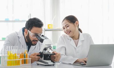 A male scientist is using a microscope to look and do some research. with a female assistant sitting near him and using Laptop or notebook to Keep a note of what he says in the laboratory.