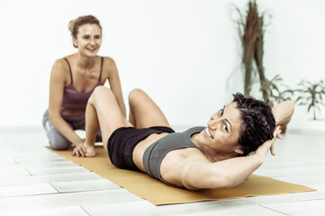 Fototapeta na wymiar Woman helping her friend woman to train abdominal muscles by twisting while lying on mat indoors. Workout together