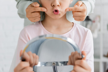 Cropped view of speech therapist pointing at cheeks while kid talking near mirror in consulting room.