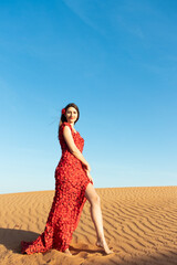 Young beautiful woman in long red dress with red rose petals among the desert. Desert rose conception.