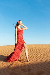 Young beautiful woman in long red dress with red rose petals among the desert. Desert rose conception.