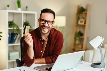 Young happy man is holding a credit card in his hand while sitting at a table with a laptop in front