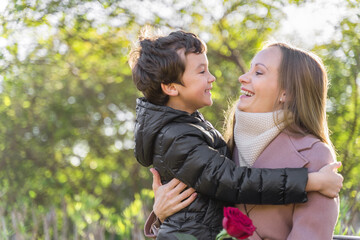 Young woman looking with love and smile at her son and hold a red pink rose. Happy boy is smiling and hugging his mother. Happy mother's day