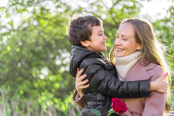 Fototapeta premium Young woman smiling, embracing a son. Happy boy is smiling and hugging his mother. Red pink rose in mother day. 