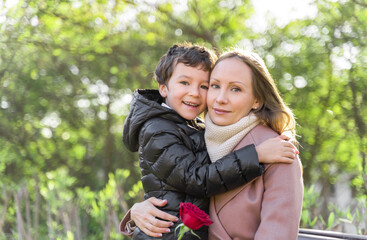 Young women and 9 years old boy hug each other and look forward into camera. Mother holding a red pink rose