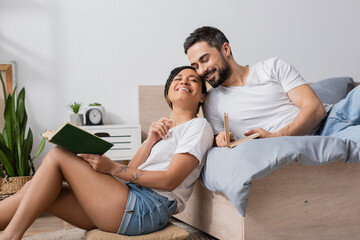 carefree african american woman with book smiling with closed eyes while sitting near smiling boyfriend lying on bed at home.