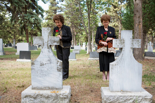 Two Women Holding Single Red Roses Standing At Two Graves Side By Side In A Cemetery; Edmonton, Alberta, Canada