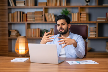 Millennial arab freelancer man in casual sitting at work desk in front of laptop, gesturing and smiling, having video call with colleagues while working from home, copy space