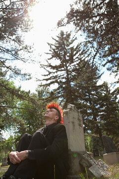 A Young Man With A Red Mohawk Sits Alone By A Tombstone In A Cemetery; Edmonton, Alberta, Canada