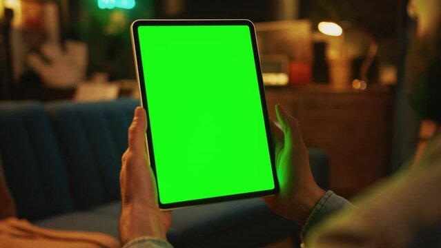 In the evening hands man holding and tapping on a tablet computer with a vertical green screen. Mock up. Sofa on background