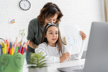 Woman kissing daughter holding paper with th letters during speech therapy video lesson at home.