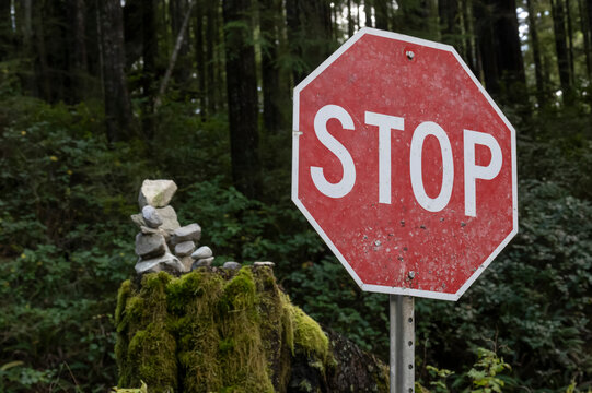 Close-up of a stop sign beside a forested area with moss and a cairn in the background in the community of Bamfield on Vancouver Island; Bamfield, British Columbia, Canada