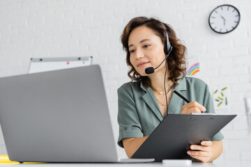 Speech therapist in headset writing on clipboard and looking at laptop in consulting room.