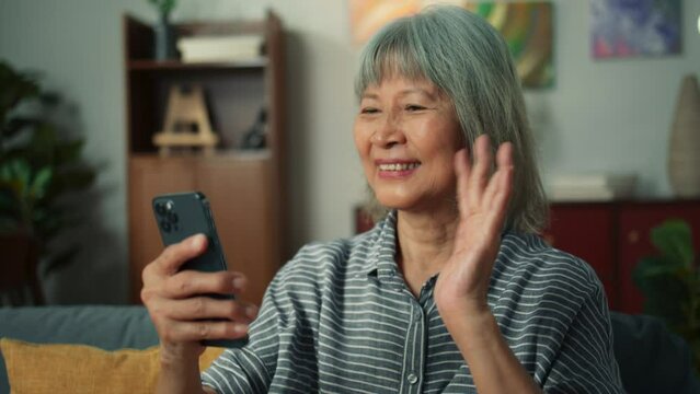 Happy attractive Asian elderly woman using smartphone video conference call in cozy living room. Happy old aged female using mobile phone technology chatting talking with family or friends at home
