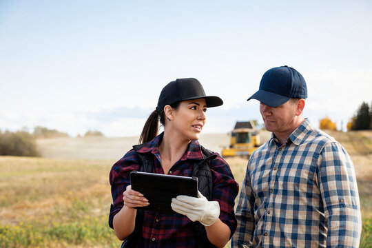 Close-up of a husband and wife using portable wireless device to manage and monitor their canola harvest with a combine harvester in the background; Alcomdale, Alberta, Canada