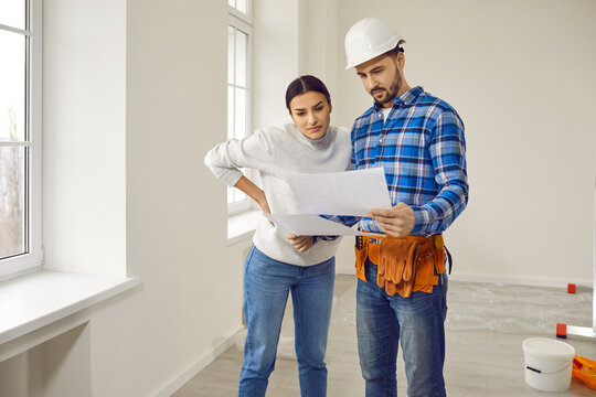 Finishing Painting Decorating Work Foreman Builder Supervisor In Uniform Hardhat Showing Young Woman Homeowner Renovation Plan In Empty House Or Apartment With Bucket Of Paint Or Plaster In Background