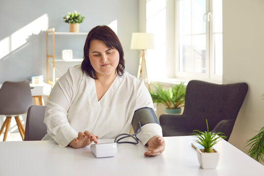 Overweight woman with hypertension symptoms does self checkup at home. Fat young girl sitting at table with cuff of digital sphygmomanometer on arm taking blood pressure and heartbeat rhythm herself