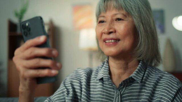 Happy attractive Asian elderly woman using smartphone video conference call in cozy living room. Happy old aged female using mobile phone technology chatting talking with family or friends at home