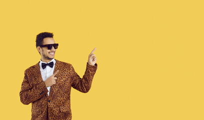 Positive man in funky outfit, bow tie and sunglasses pointing finger hand gesture to side at free blank advertising copyspace studio background, promoting novelty, suggesting something new at discount