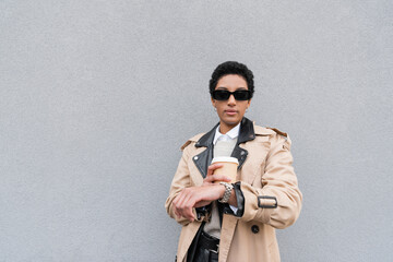african american businesswoman in sunglasses holding paper cup and checking time on wristwatch while waiting on street.