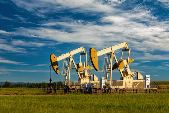 Two pumpjacks in a green field reflecting the warm light at sunrise with dramatic clouds and blue sky in the background, West of Airdrie; Alberta, Canada