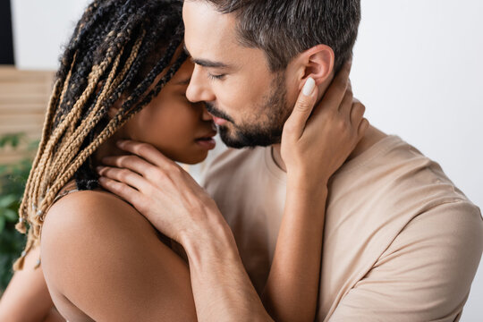 side view of brunette bearded man and hot african american woman with dreadlocks kissing at home.