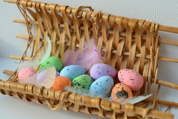  Easter eggs and colorful feathers in a wicker basket on a light background. Easter