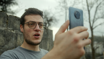 Shocked Handsome Young Man with Glasses Having a Video Call on Smartphone Outdoors