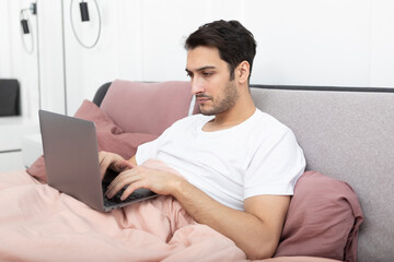 Handsome man lying on the bed and working on laptop in the bedroom	