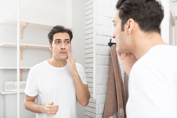 Obraz premium Handsome man looking at mirror and applying moisturizing cream on cheeks in bathroom. Groomed young guy doing skincare morning routine after taking a shower