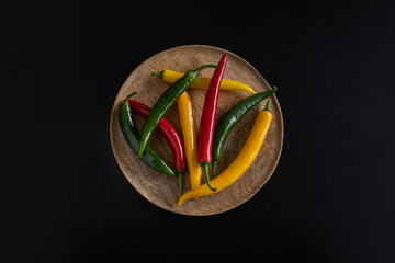 Multicolored pods of hot chili peppers lie on a wooden textured plate tray bowl on a black background with copy space