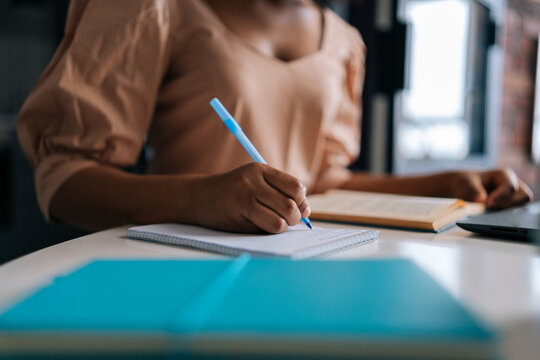 Detail cropped shot of unrecognizable African American female student using laptop and paper book writing notes sitting at desk. Closeup black woman making notes studying prepare essay