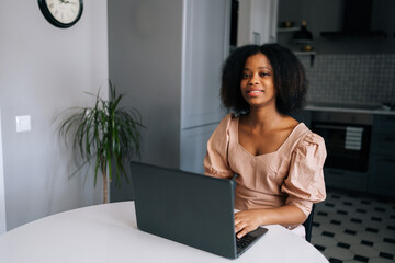 Medium shot of friendly African young woman sitting at table in modern kitchen room working on laptop, smiling looking at camera. Positive black female freelancer doing online job from home office.