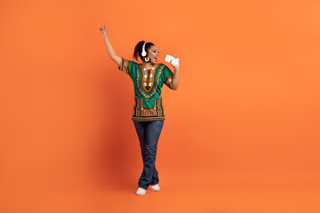 Cheery black lady in african clothing singing, dancing on orange