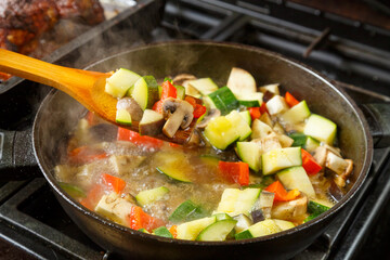 Frying pan with stewed vegetables in sauce and spices and a wooden spoon on the stove .