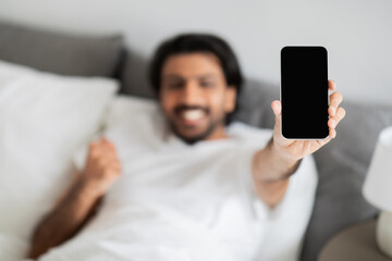 Cheerful young middle eastern man in white t-shirt lies on bed, makes success gesture, celebrates victory