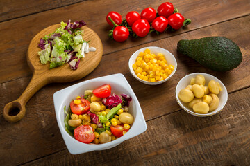 Vegetable salad with lettuce and cherry tomato with avocado olive oil in a white plate on a wooden table next to all the ingredients.