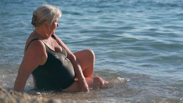 Elderly senior woman with gray hair, sitting in shallow calm sea near beach, enjoying sunshine