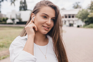 Relaxed young woman brunette in wireless headphones walk on the street listening to music. Enjoying listening to lounge music live via smartphone app, relaxed, eyes closed, laughing, touch earphones.
