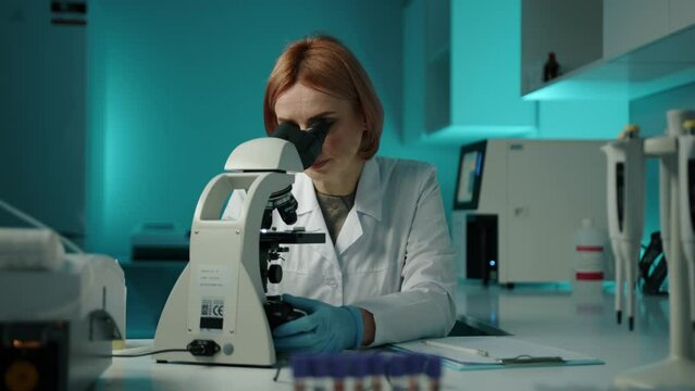 A veterinarian in a white lab coat looking at the sample through a microscope. She is in a laboratory and is wearing glasses. High quality 4k footage 