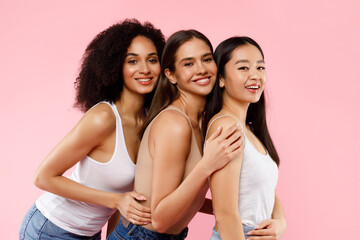 It girls. Happy three multiracial young ladies in casual embracing, posing and smiling at camera on pink background