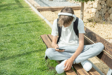 Teenage boy sitting outdoors and using smart phone to listen music with headphones, watching video, writing sms, scrolling news, playing games, chating with friends, using messaging or video chat app