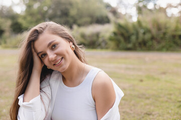 Nice looking woman is sitting on the grass in park, hold her head and dreaming. She is relaxing, enjoying of the day. Girl look sensual and tenderness. Long hair female wear white shirt and top.