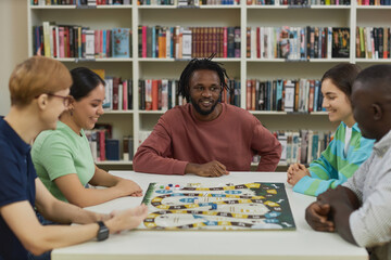 Diverse group of young people playing board games together while sitting around table in library...