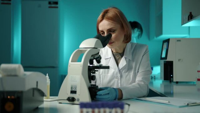 A woman in a white lab coat writing down something down and looking through a microscope. She is in a laboratory and is wearing glasses High quality 4k footage 
