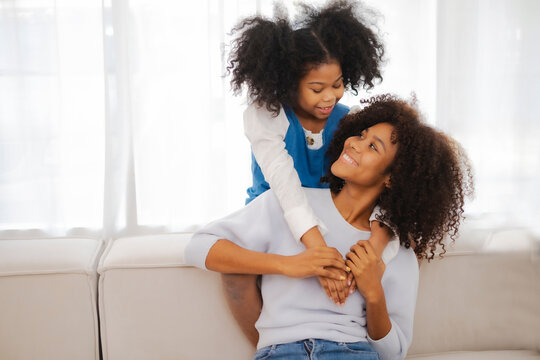African American kid sitting over sofa embrass her mother neck, look in her eye with love feeling, while mom look back with smile, expression of love in mother's day.