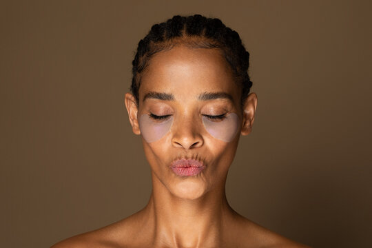 Black middle aged woman with hydrogel patches under eye blowing kiss, posing isolated over brown background